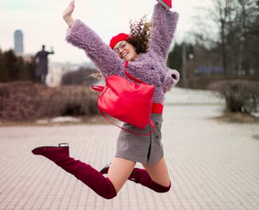 A fashionable young woman jumping with joy outdoors, wearing vibrant clothing and accessories.